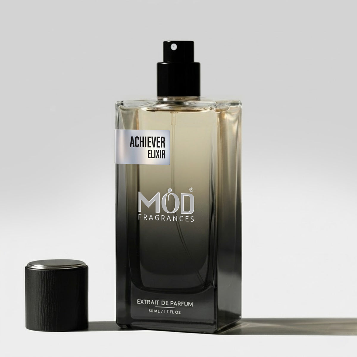 A rectangular glass perfume bottle by MOD Fragrances, labeled "Achiever Elixir” stands on a light surface with its black cap placed beside it.
