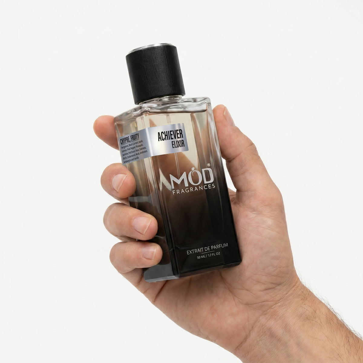 A Male person's hand holds a rectangular glass perfume bottle by MOD Fragrances, labeled "Achiever Elixir” against a plain white background.