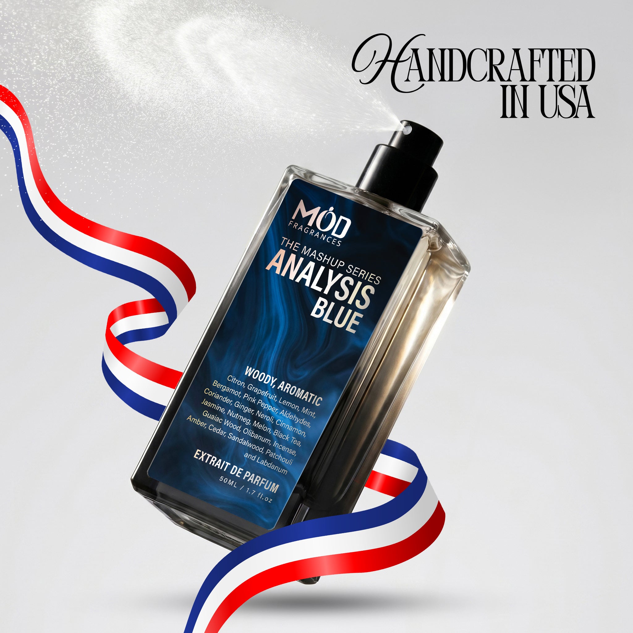 A rectangular glass perfume bottle by MOD Fragrances, labeled "Mashup series Analysis Blue" is captured mid-spray with a fine mist dispersing into the air, accented by stylized red, white, and blue ribbons, with the text "Handcrafted in USA" in the upper right.