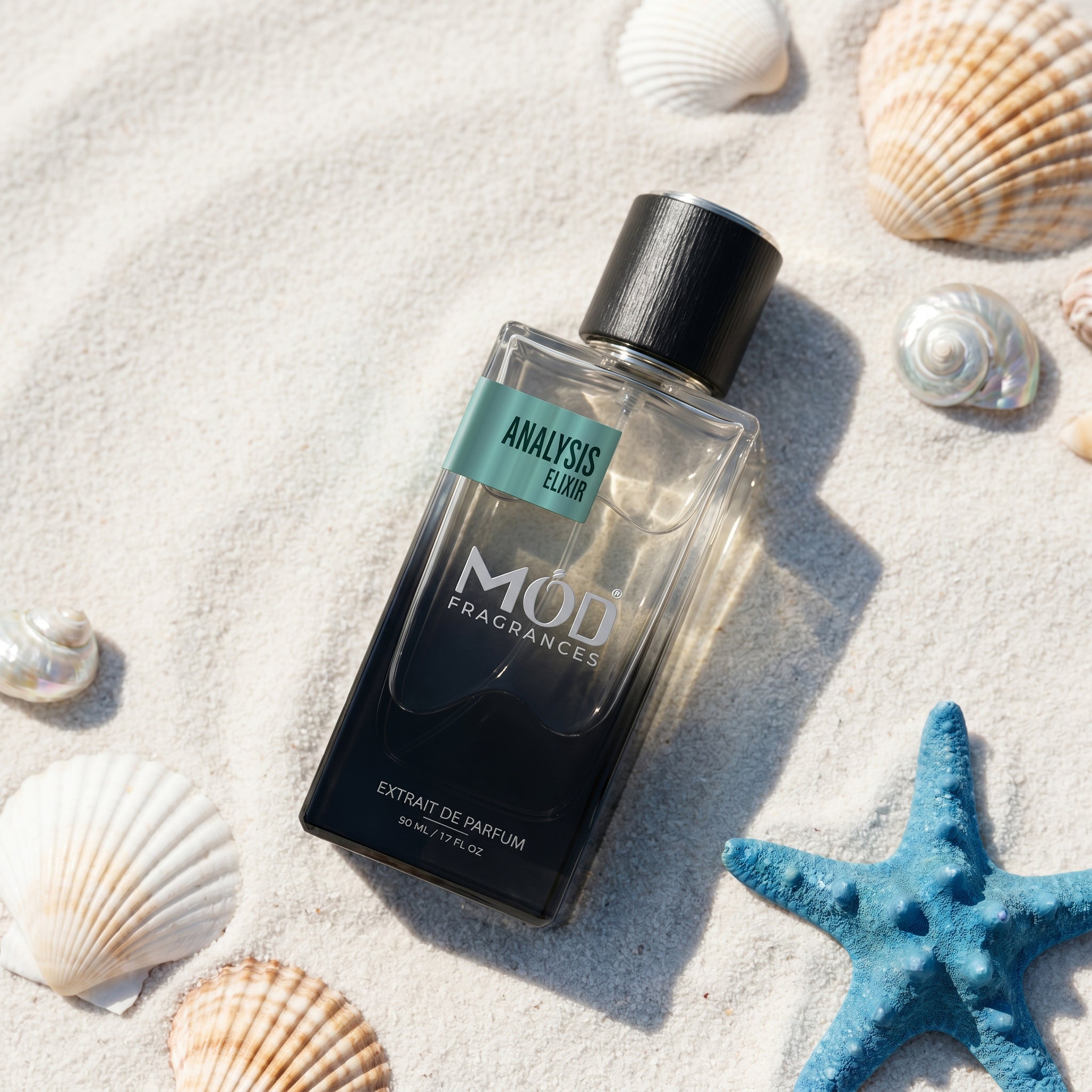 A rectangular glass perfume bottle by MOD Fragrances, labeled "Analysis Elixir," is placed on light-colored sand and surrounded by various seashells and a blue starfish.