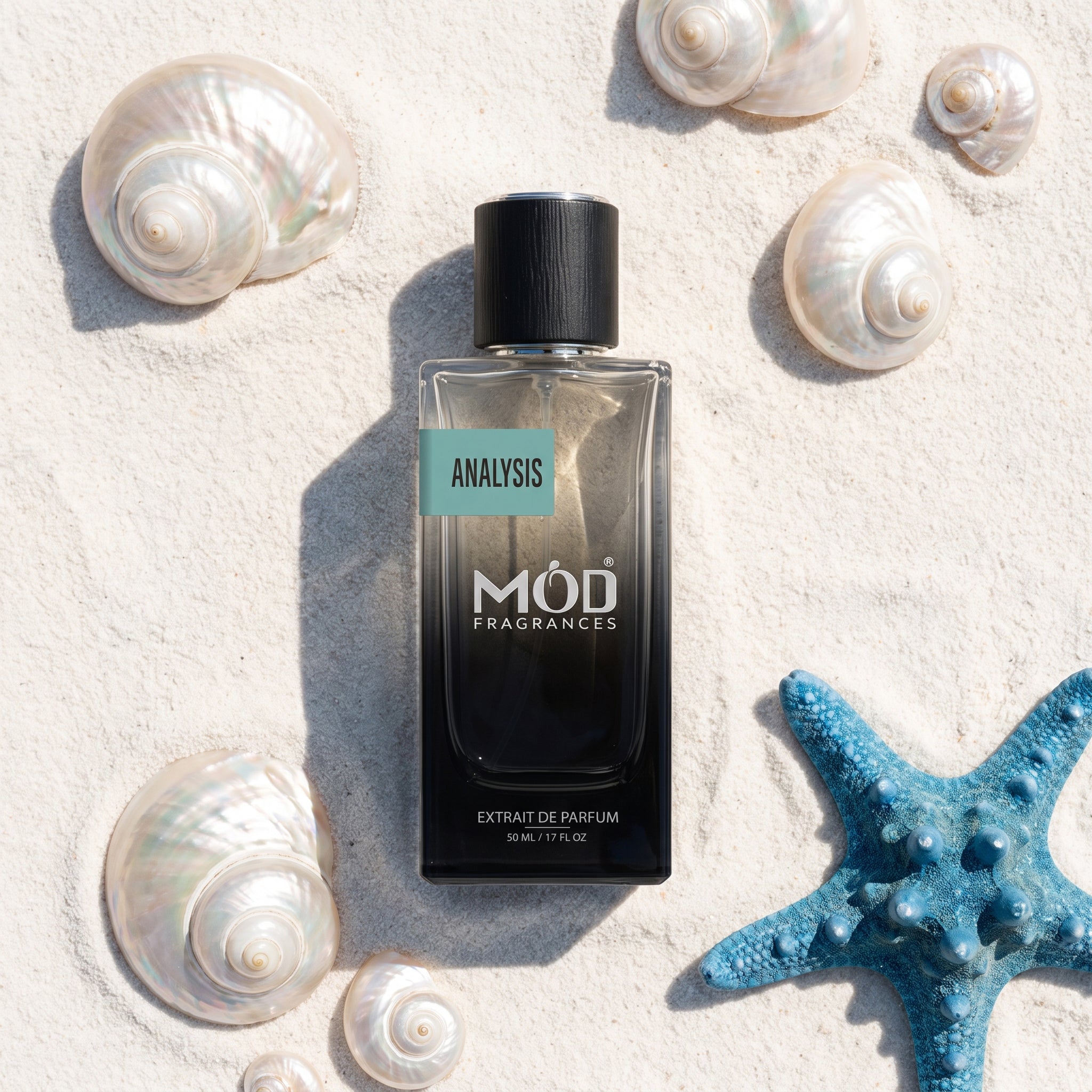 A rectangular glass perfume bottle by MOD Fragrances, labeled "Analysis" is placed on light-colored sand and surrounded by various seashells and a blue starfish.