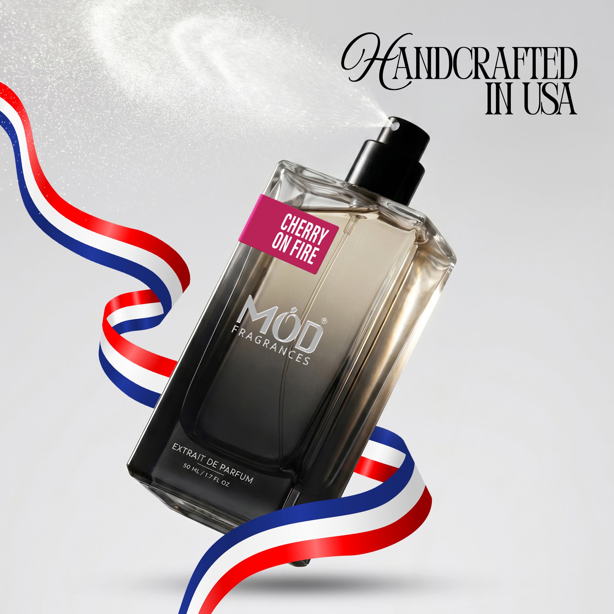A rectangular glass perfume bottle by MOD Fragrances, labeled "Cherry on Fire" is captured mid-spray with a fine mist dispersing into the air, accented by stylized red, white, and blue ribbons, with the text "Handcrafted in USA" in the upper right.