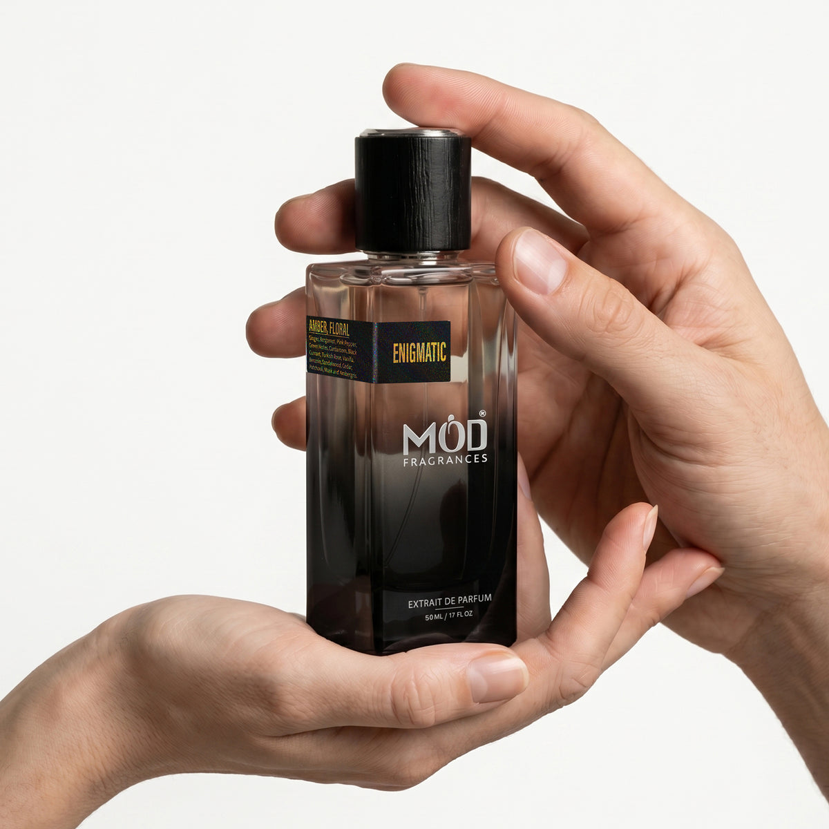 A Male and a female person's hand holds a rectangular glass perfume bottle by MOD Fragrances, labeled "Enigmatic” against a plain white background.