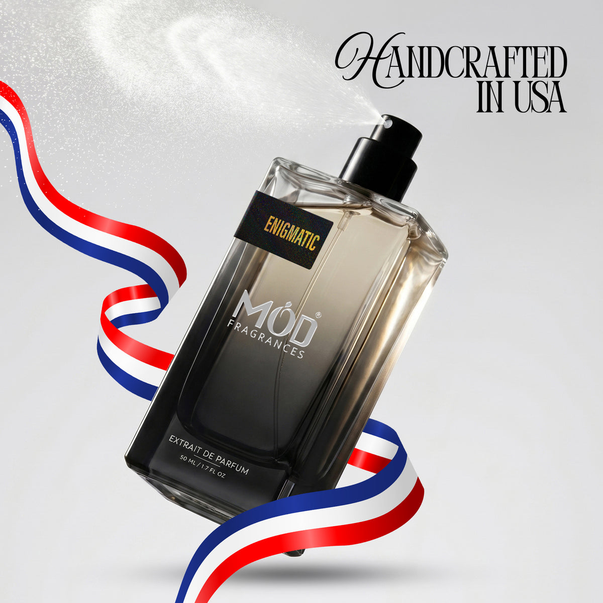 A rectangular glass perfume bottle by MOD Fragrances, labeled "Enigmatic” is captured mid-spray with a fine mist dispersing into the air, accented by stylized red, white, and blue ribbons, with the text "Handcrafted in USA" in the upper right.