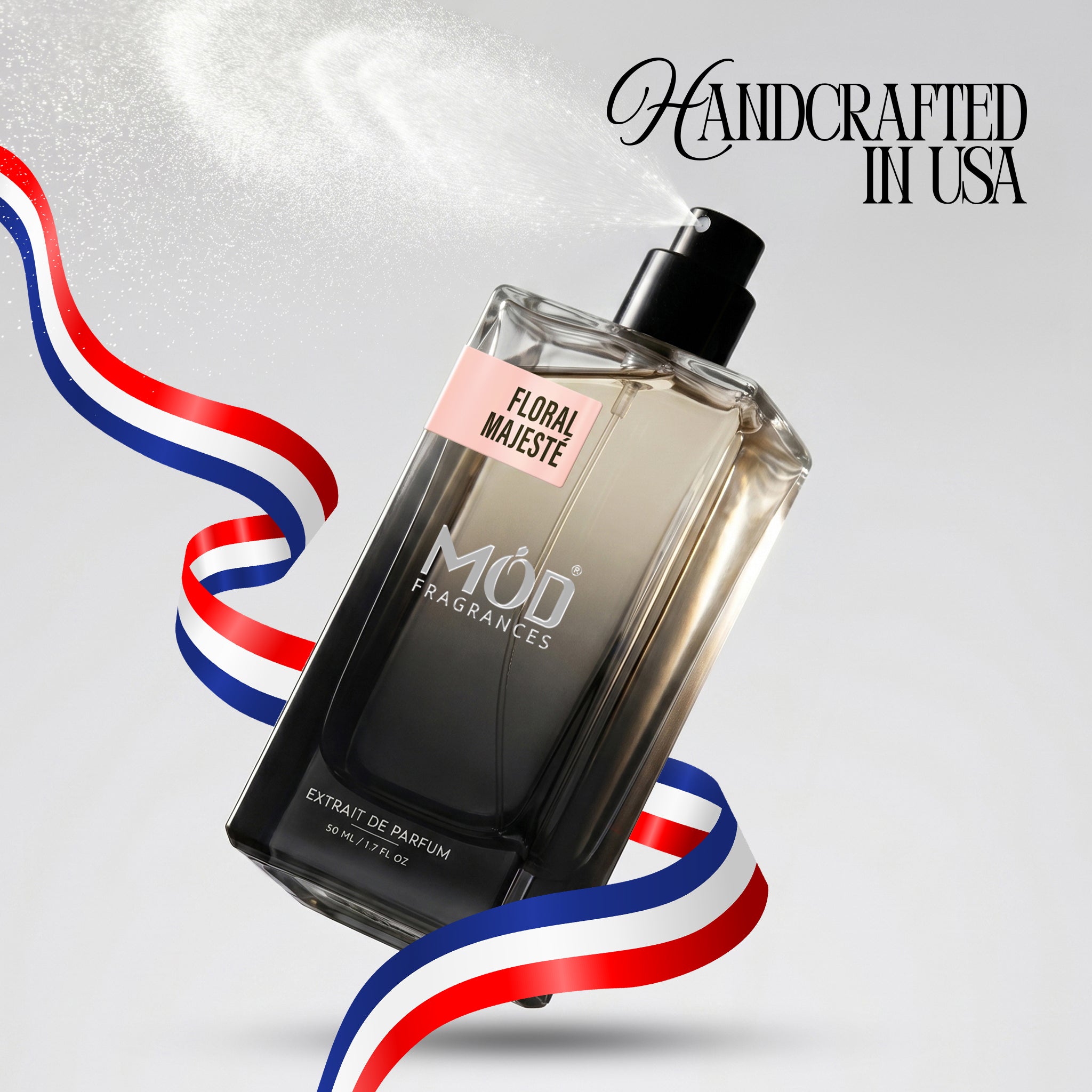 A rectangular glass perfume bottle by MOD Fragrances, labeled "Floral Majeste” is captured mid-spray with a fine mist dispersing into the air, accented by stylized red, white, and blue ribbons, with the text "Handcrafted in USA" in the upper right.
