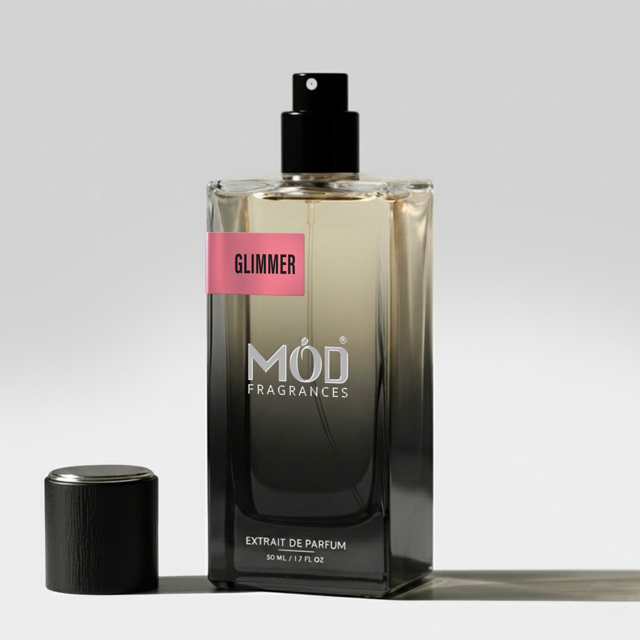 A rectangular glass perfume bottle by MOD Fragrances, labeled "Glimmer" stands on a light surface with its black cap placed beside it.