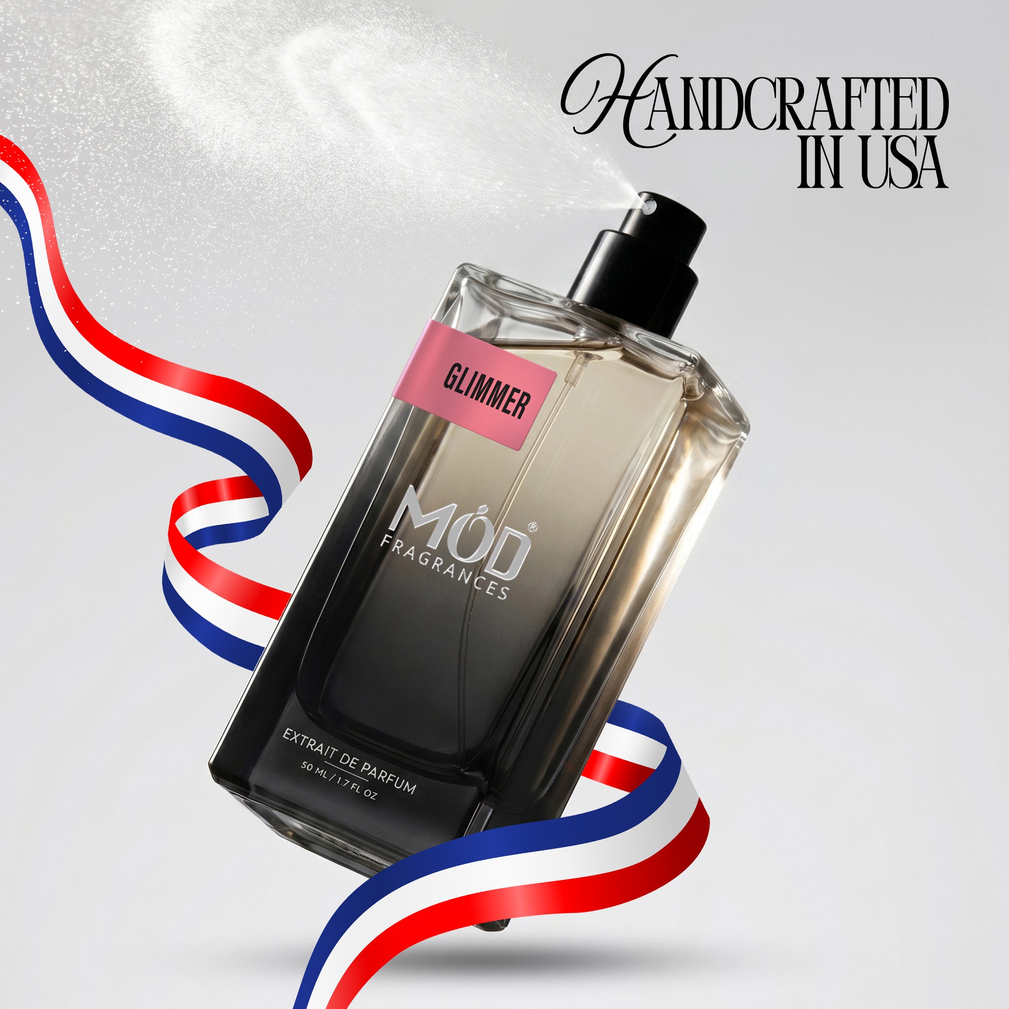 A rectangular glass perfume bottle by MOD Fragrances, labeled "Glimmer" is captured mid-spray with a fine mist dispersing into the air, accented by stylized red, white, and blue ribbons, with the text "Handcrafted in USA" in the upper right.