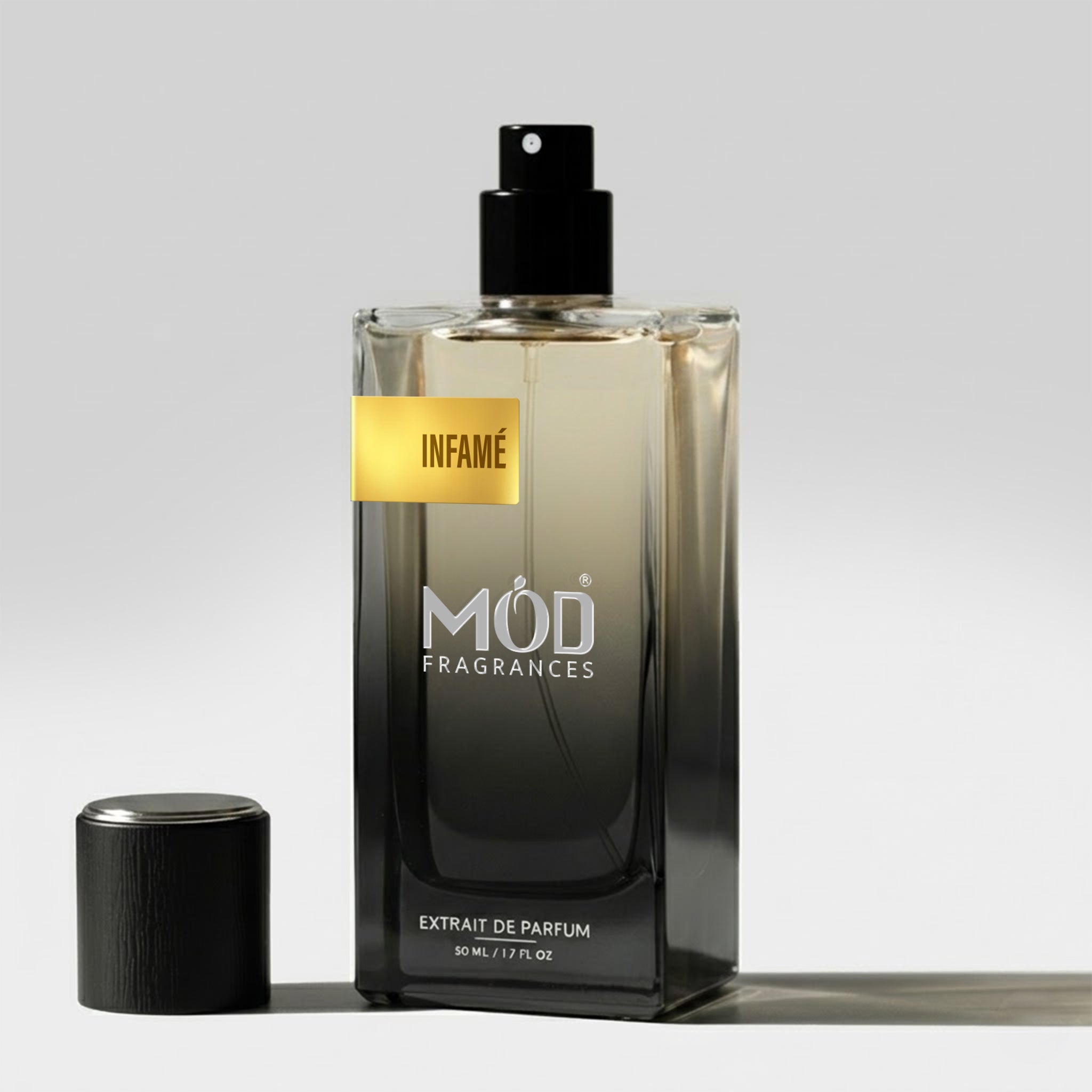 A rectangular glass perfume bottle by MOD Fragrances, labeled "Infame” stands on a light surface with its black cap placed beside it.