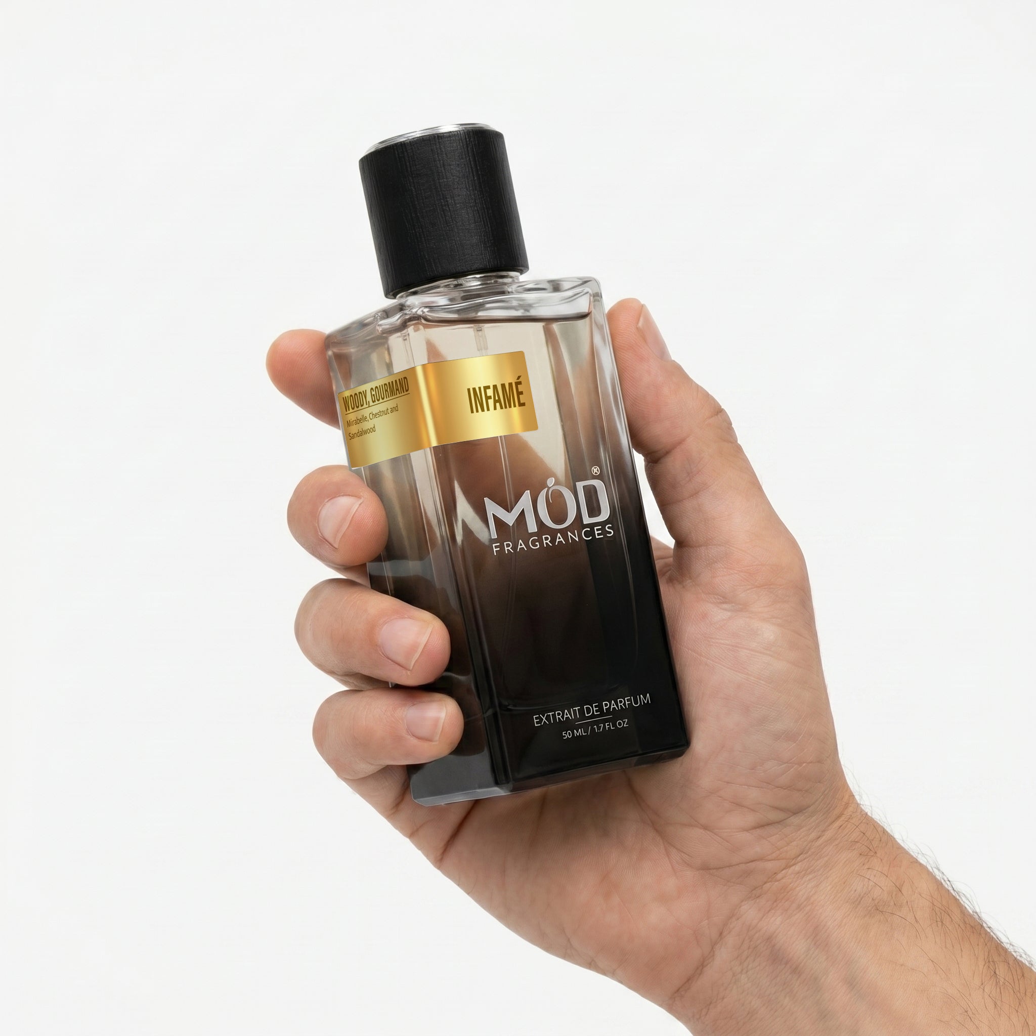 A Male person's hand holds a rectangular glass perfume bottle by MOD Fragrances, labeled "Infame” against a plain white background.