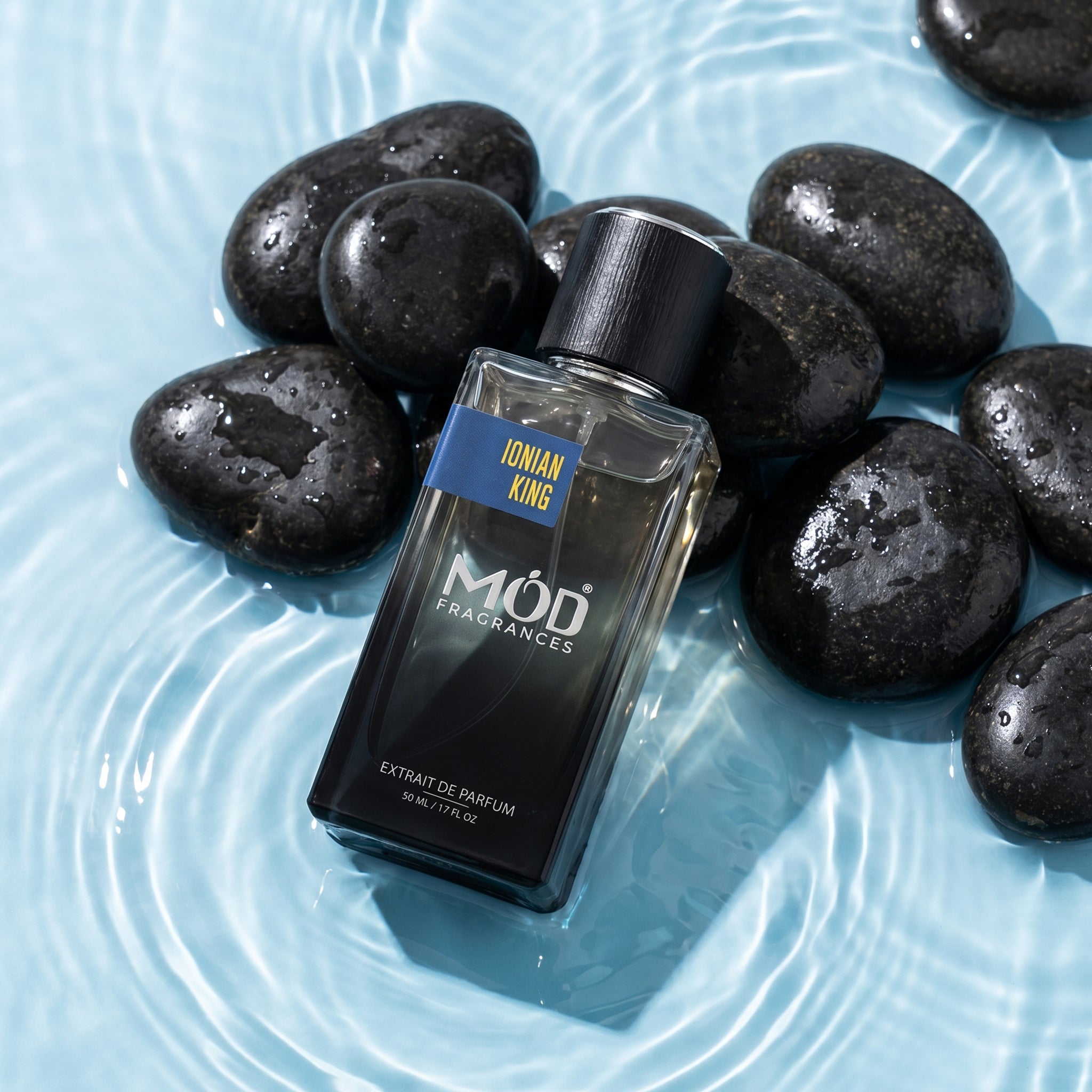 A rectangular glass perfume bottle by MOD Fragrances, labeled "Ionian King" is placed on black round and oval pebbles, half merged in clear shallow water, 