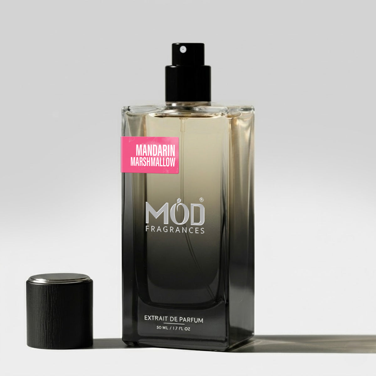 A bottle of MOD Fragrances Extrait de Parfum in the Mandarin Marshmallow scent, featuring a black gradient bottle with a pink label, shown next to its black cap on a light gray background.
