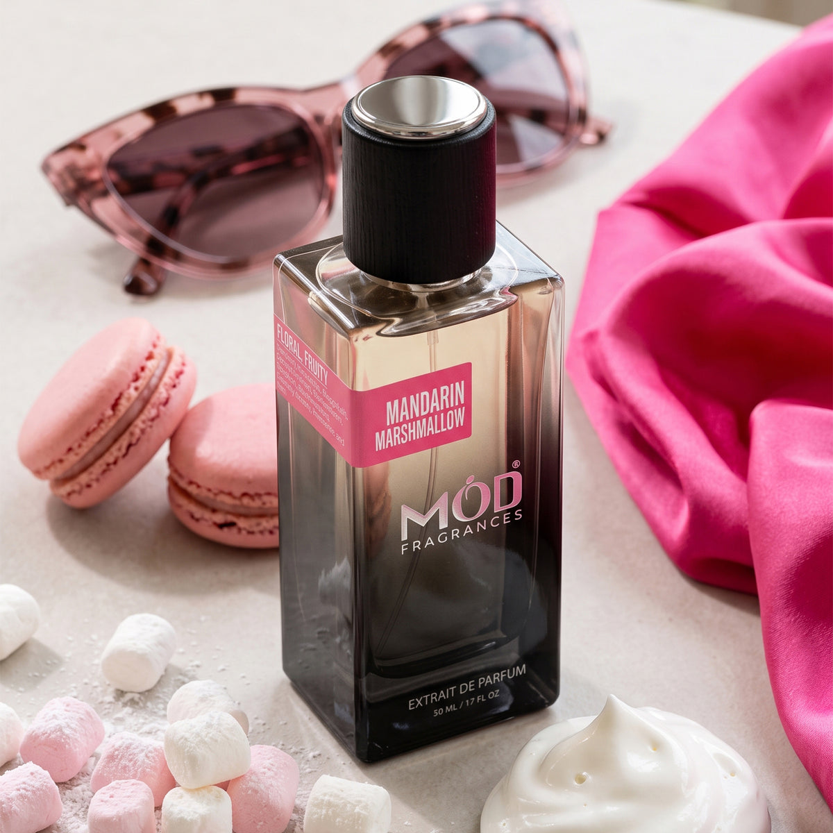 A bottle of MOD Fragrances Mandarin Marshmallow extrait de parfum stands on a light surface with pink macarons, sunglasses, a pink fabric cloth, marshmallows, and whipped cream around it.