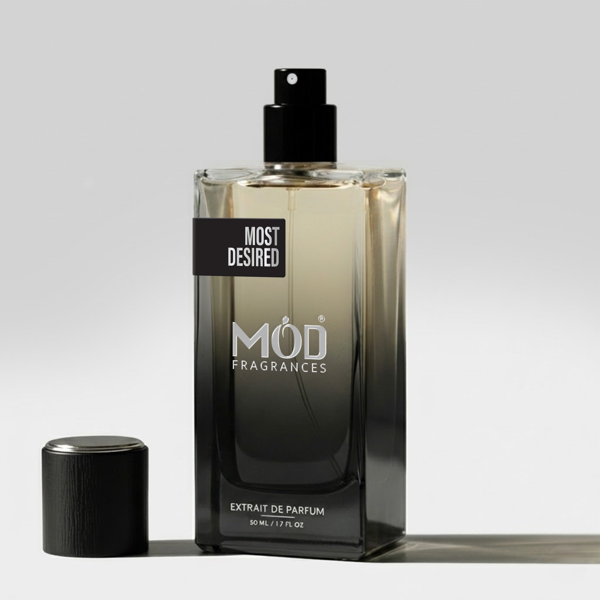A rectangular glass perfume bottle by MOD Fragrances, labeled "Most Desired" stands on a light surface with its black cap placed beside it.