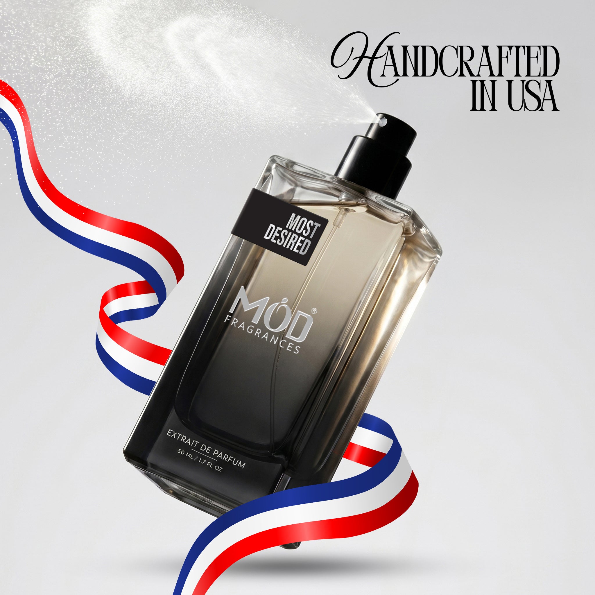 A rectangular glass perfume bottle by MOD Fragrances, labeled "Most Desired" is captured mid-spray with a fine mist dispersing into the air, accented by stylized red, white, and blue ribbons, with the text "Handcrafted in USA" in the upper right.