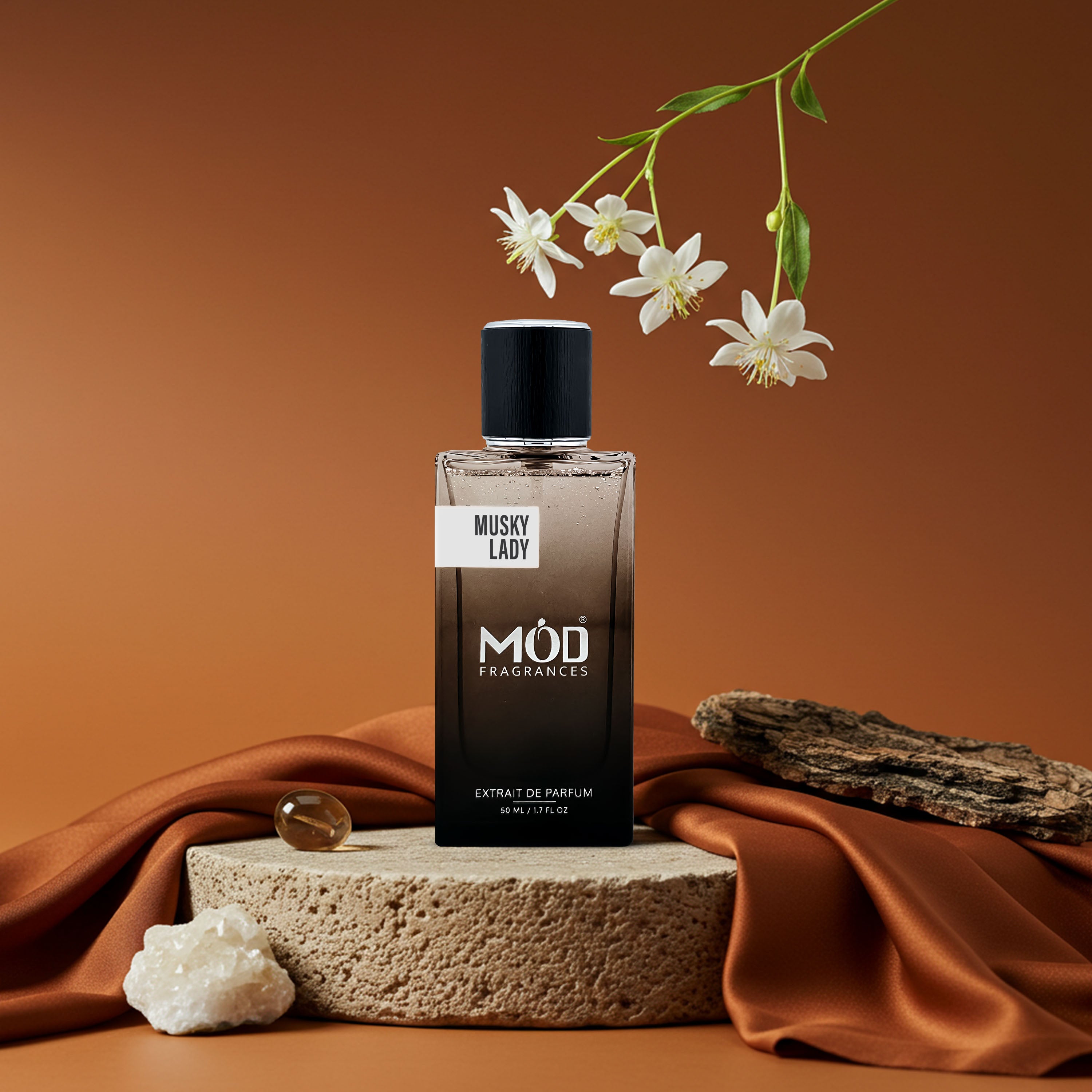 A bottle of MOD Parfums 'Musky Lady' extract de parfum stands on a textured stone pedestal draped with rust-colored satin fabric, with white flowers and a piece of driftwood in a warm brown studio setting.