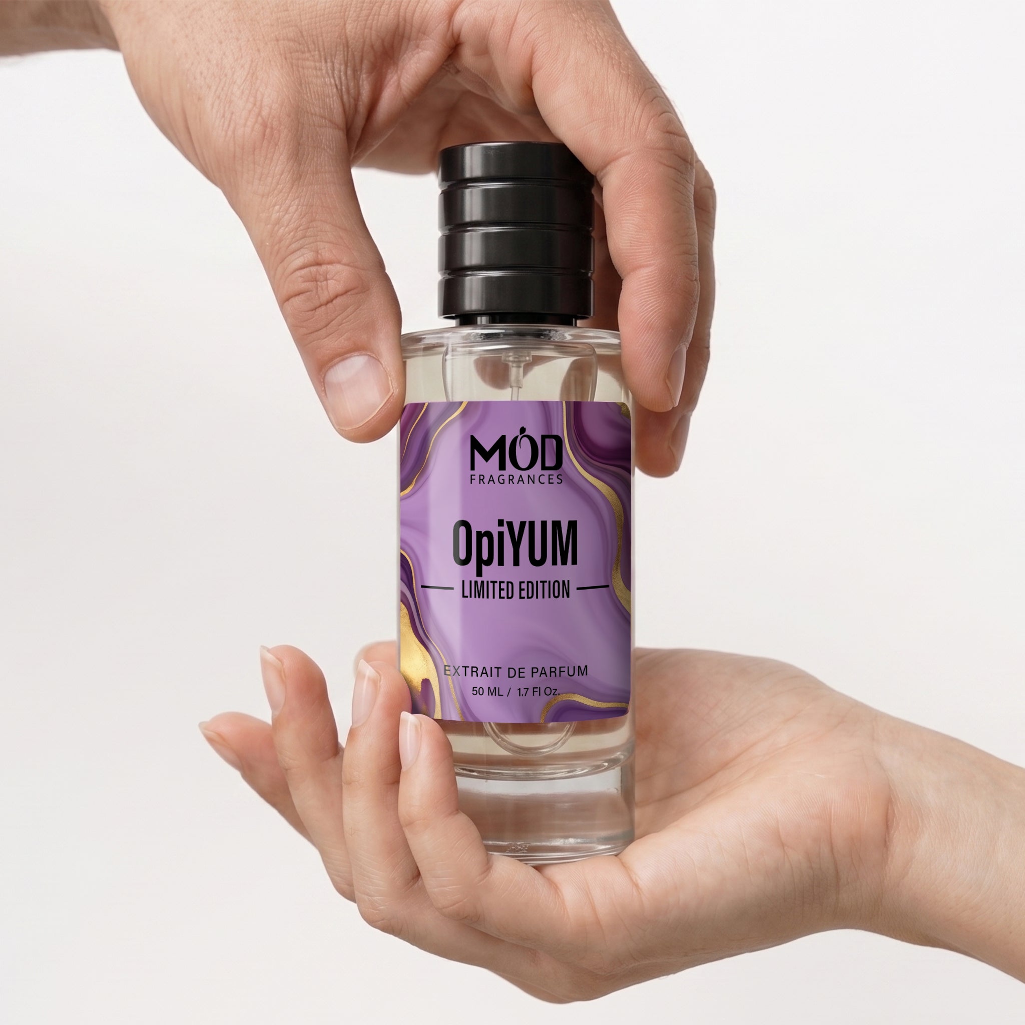 A hand holding a clear glass bottle of OpiiYUM Limited Edition Extrait de Parfum by MóD Fragrances, with another hand supporting the base. The bottle has a black ribbed cap and a purple label with gold accents.