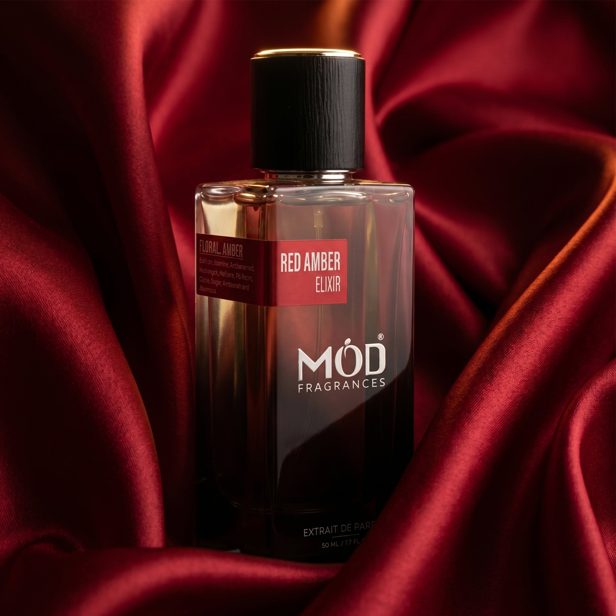 A rectangular glass perfume bottle by MOD Fragrances, labeled "Red Amber Elixir" is nestled inside red silk satin fabric