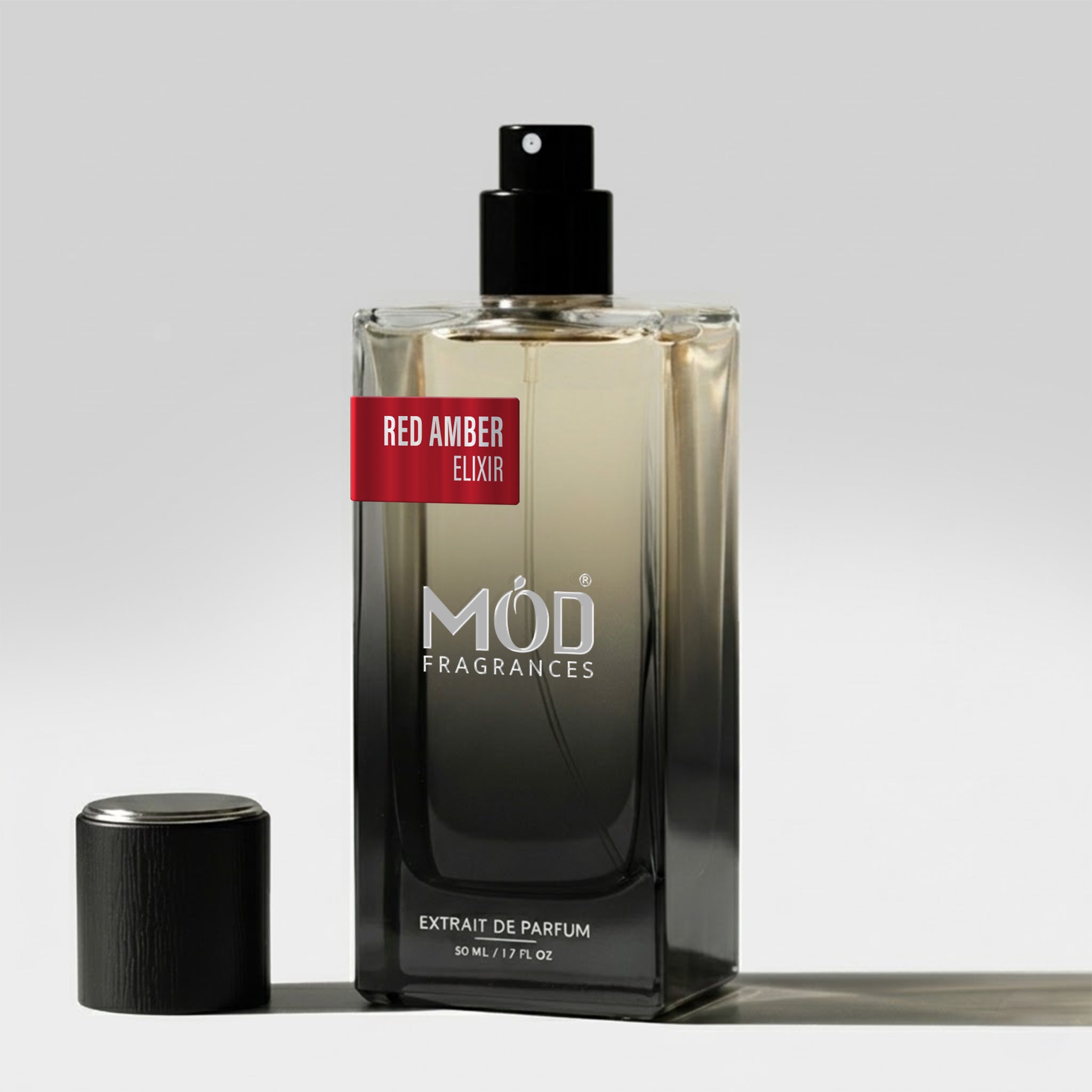 A rectangular glass perfume bottle by MOD Fragrances, labeled "Red Amber Elixir" stands on a light surface with its black cap placed beside it. 