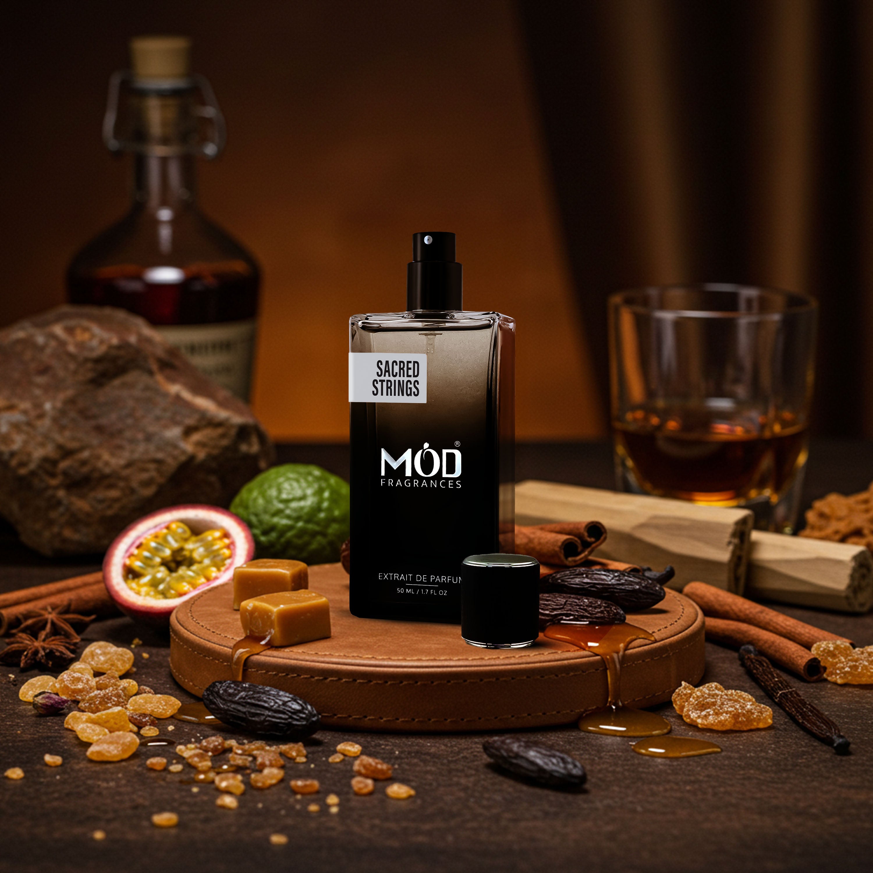 A bottle of MOD Fragrances Sacred Strings Eau de Parfum stands on a round wooden tray, surrounded by its scent ingredients including cinnamon sticks, dates, a passion fruit, and amber syrup, with a glass of whiskey and a bottle in the warm, dark background.