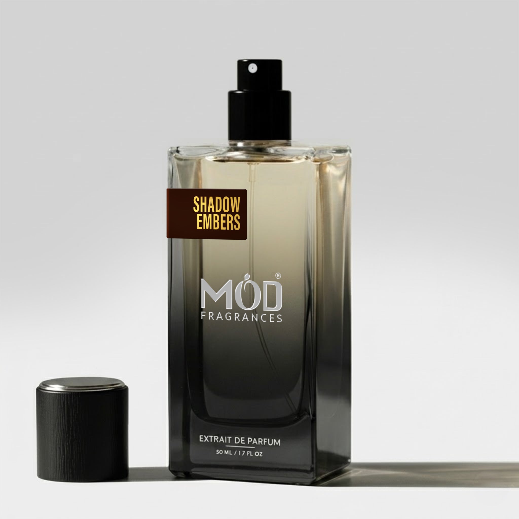 A rectangular glass perfume bottle by MOD Fragrances, labeled "Shadow Embers" stands on a light surface with its black cap placed beside it.
