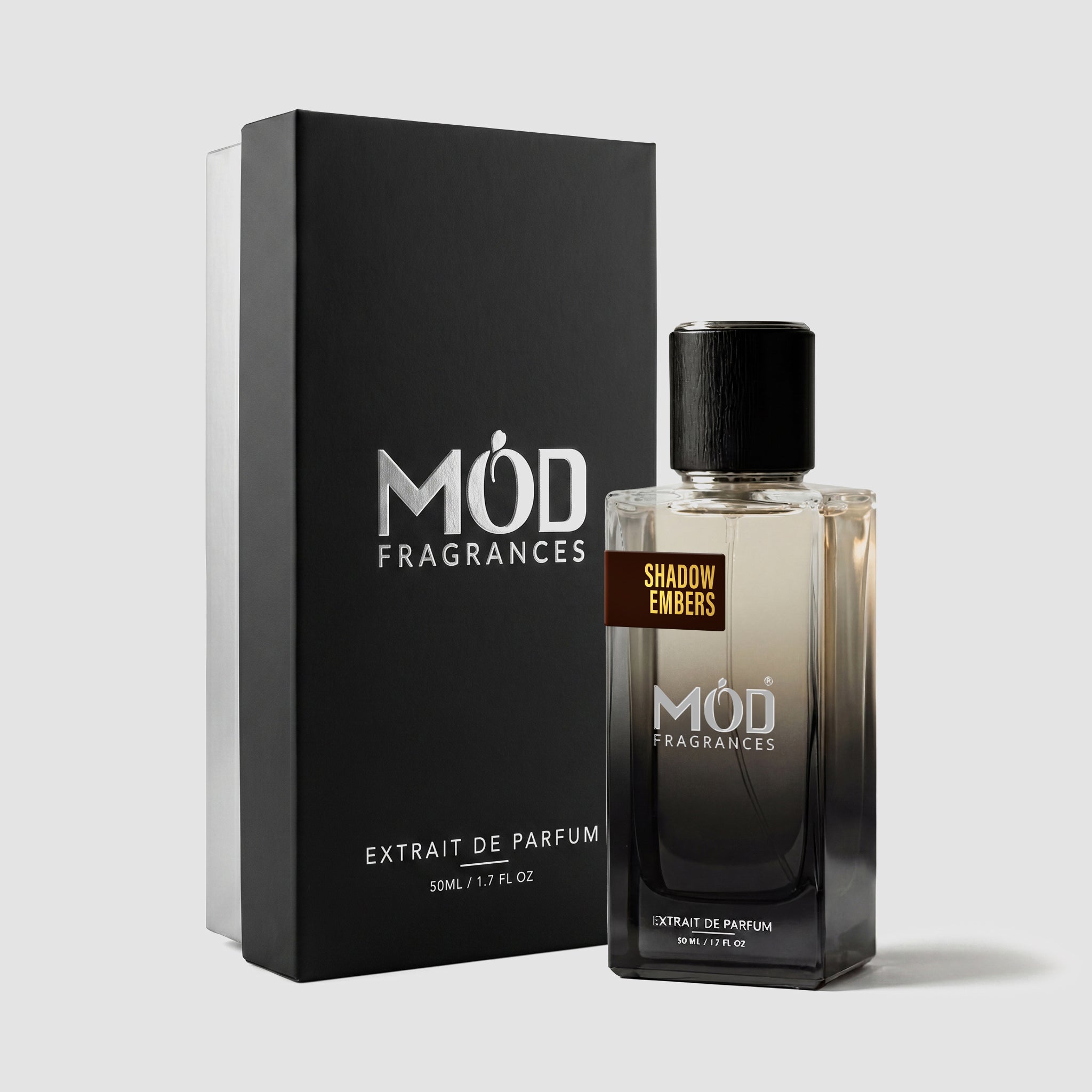 A rectangular perfume bottle by MOD Fragrances, labeled “Shadow Embers” is positioned next to its closed, upright premium black display box on a light grey background.