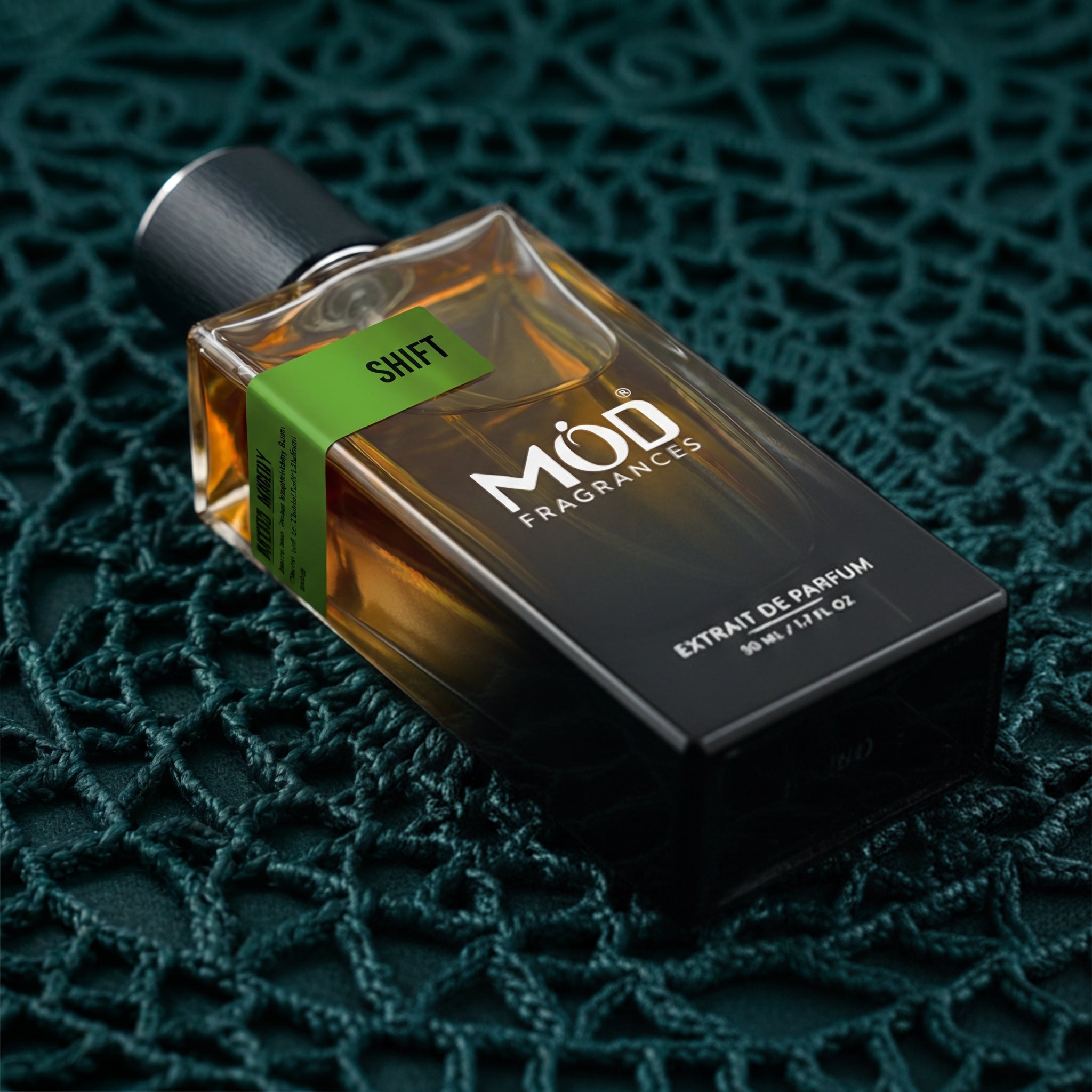 A rectangular glass perfume bottle by MOD Fragrances, labeled "Shift" is placed on dark green spiral paradigm crochet woven net,