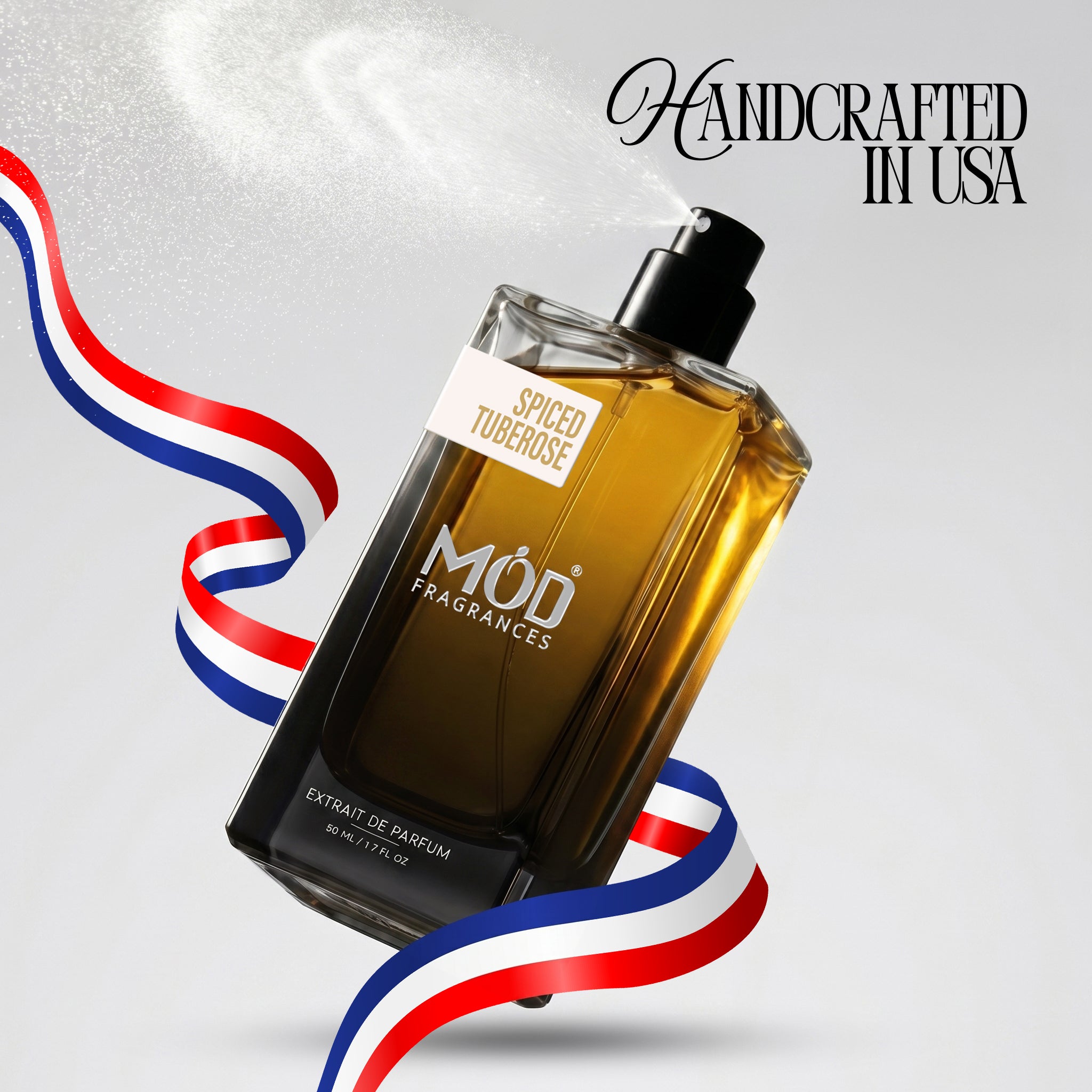 A rectangular glass perfume bottle by MOD Fragrances, labeled "Spiced Tuberose" is captured mid-spray with a fine mist dispersing into the air, accented by stylized red, white, and blue ribbons, with the text "Handcrafted in USA" in the upper right. 