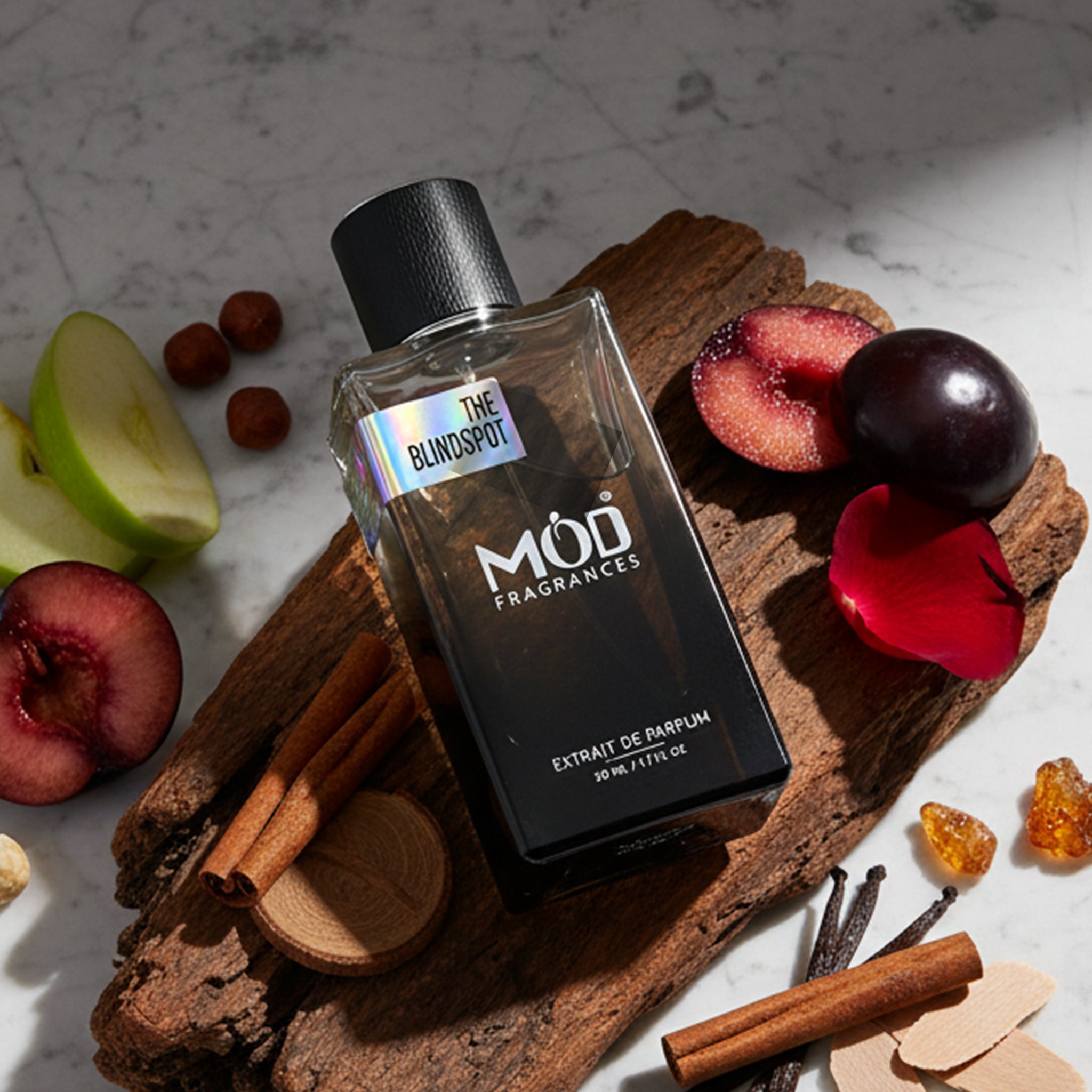 A rectangular glass perfume bottle by MOD Fragrances, labeled "The Blindspot" is placed on light-colored grey marble and large wooden bark. beside various ingredients like plum. nougat, cinnamon, green apple, amber and vanilla