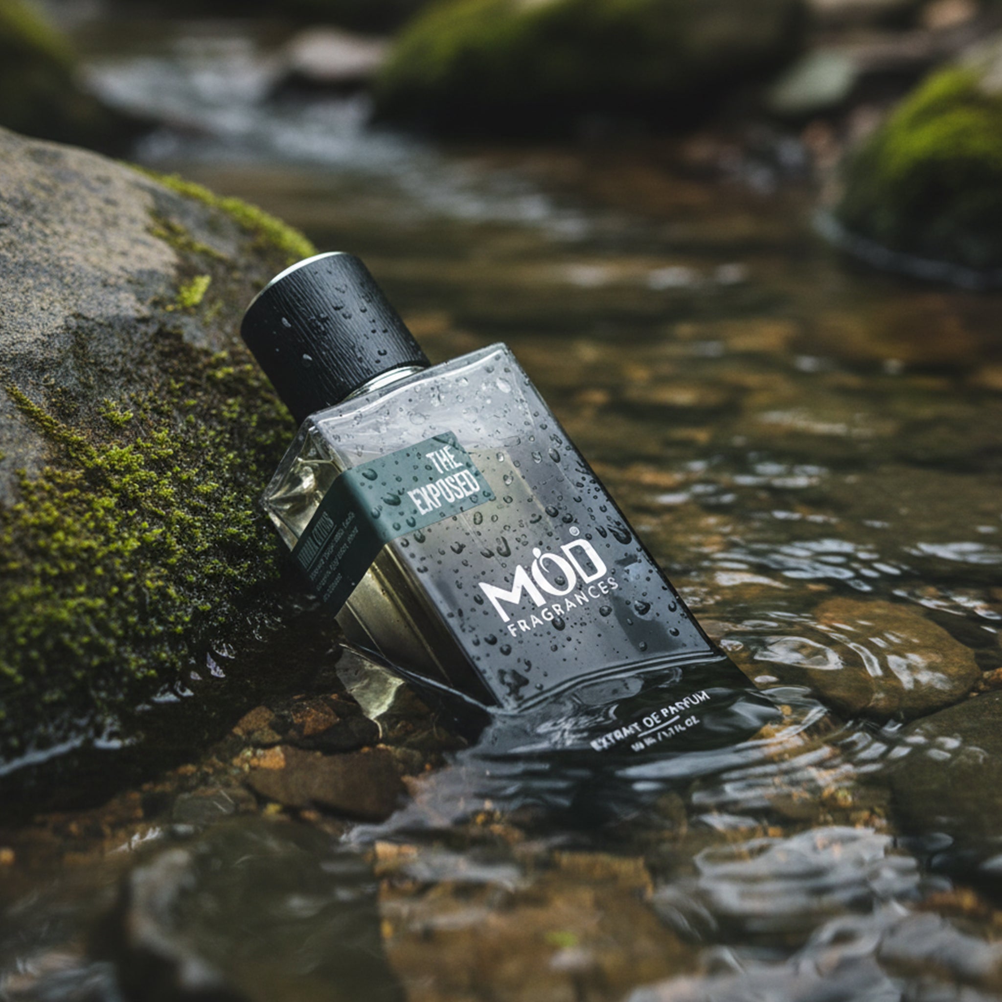 A rectangular glass perfume bottle by MOD Fragrances, labeled "The Exposed" is placed on black mossy stones, half merged in clear shallow water stream,  