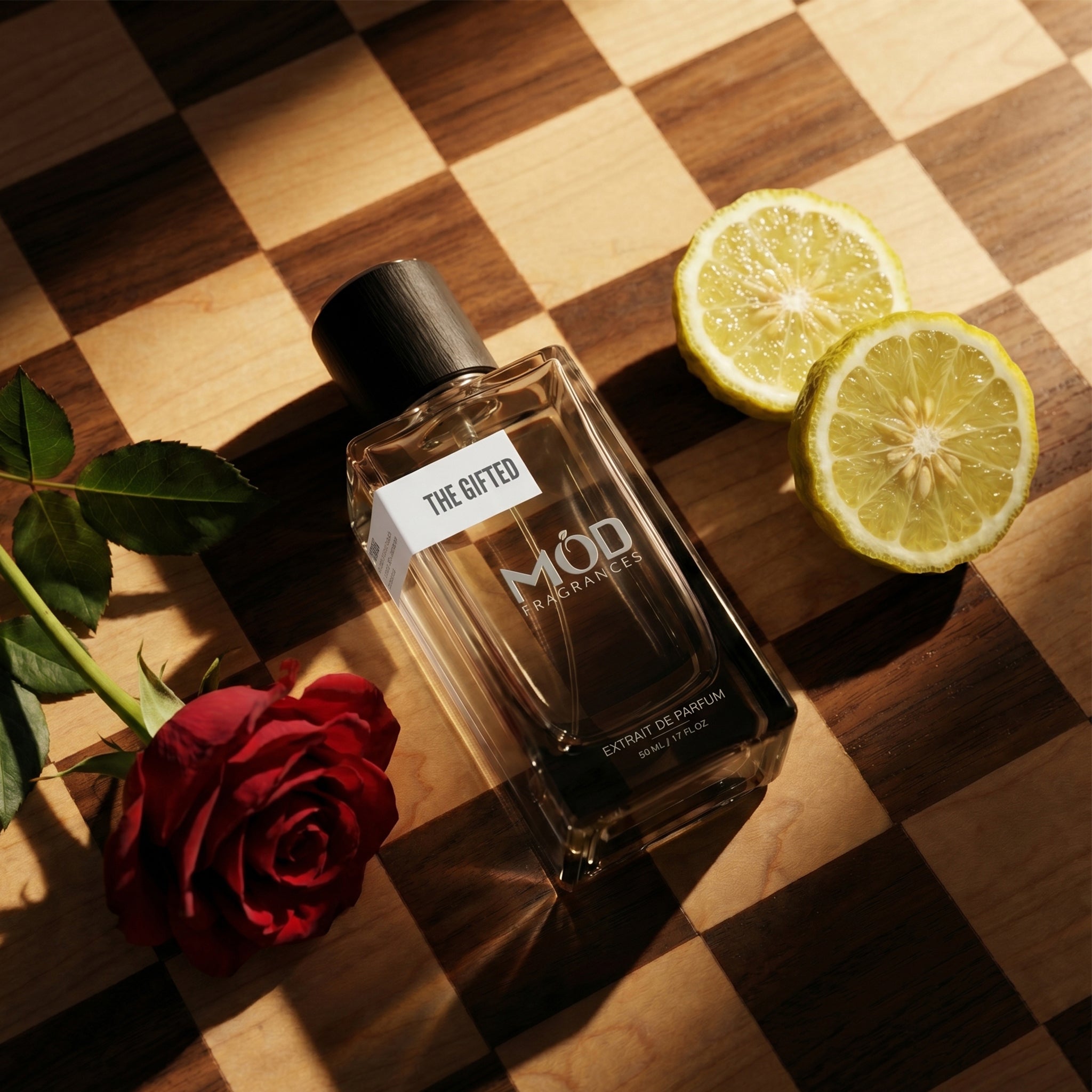 Perfume bottle labeled 'The Gifted' with a red rose and lemon slices on a checkered wooden surface