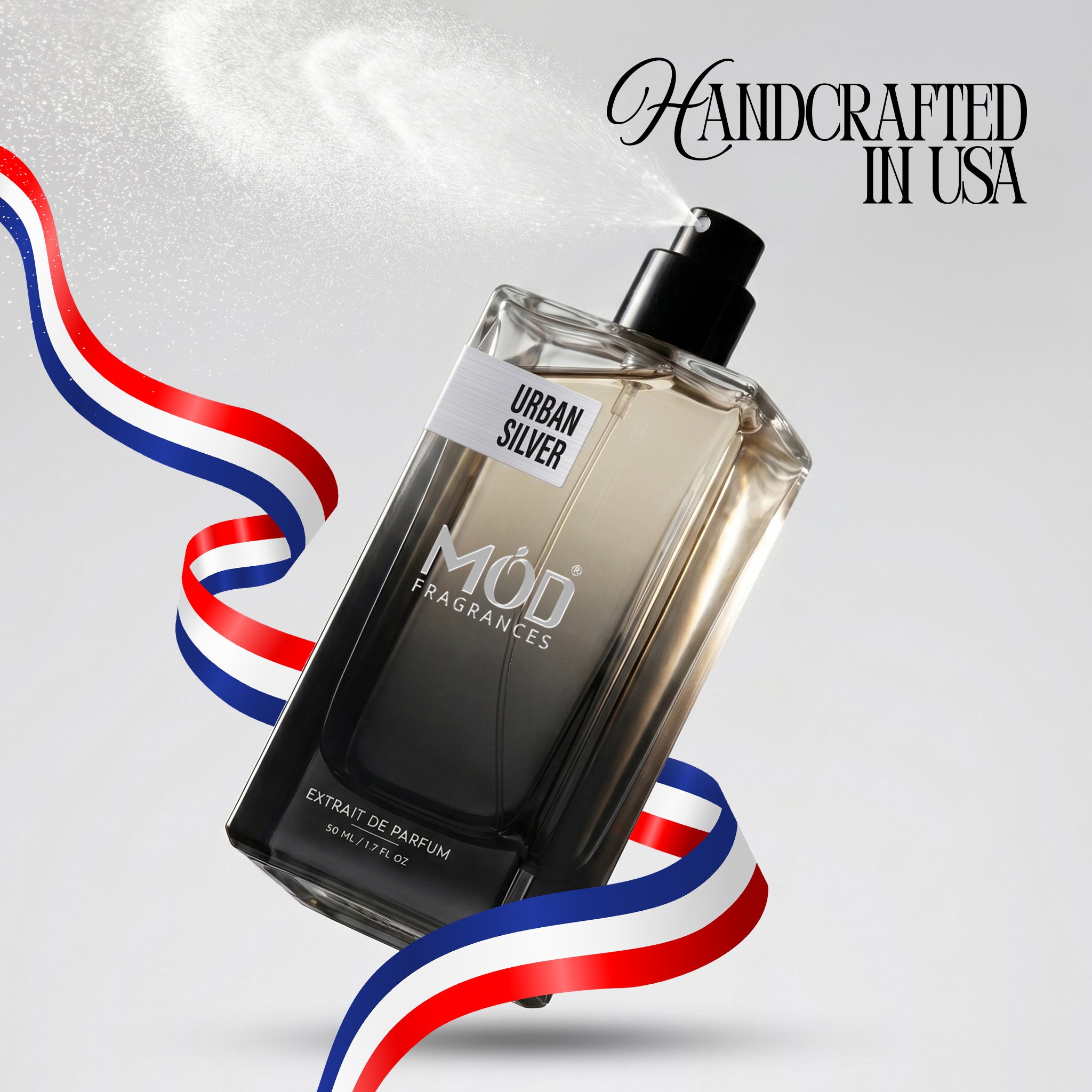 A rectangular glass perfume bottle by MOD Fragrances, labeled "Urban Silver" is captured mid-spray with a fine mist dispersing into the air, accented by stylized red, white, and blue ribbons, with the text "Handcrafted in USA" in the upper right.
