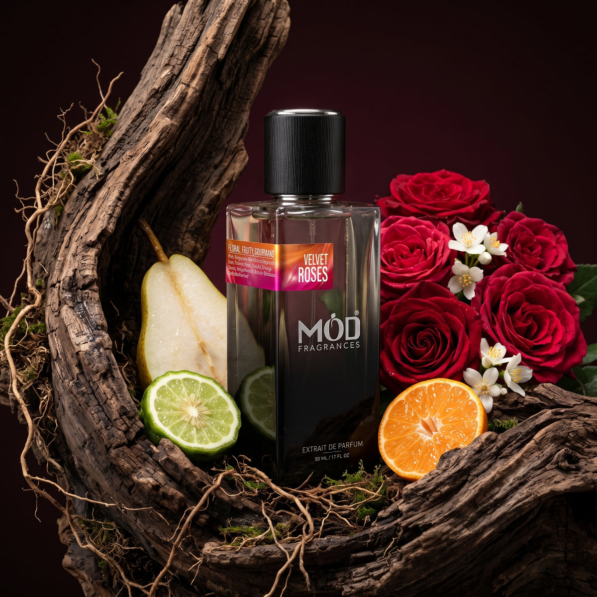 A bottle of MÓD Fragrances Velvet Roses perfume is displayed on a rustic arrangement of driftwood, surrounded by fresh pear slices, lime halves, an orange slice, and red roses against a dark burgundy background.